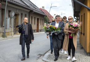 Grupa ludzi idzie po chodniku. Mężczyzna i kobieta z przodu trzymają kwiaty.