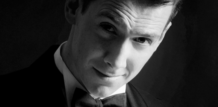 młody mężczyzna w garniturze i muszce uśmiecha się lekko patrząc na wprost, głowa delikatnie pochylona w jedną stronę