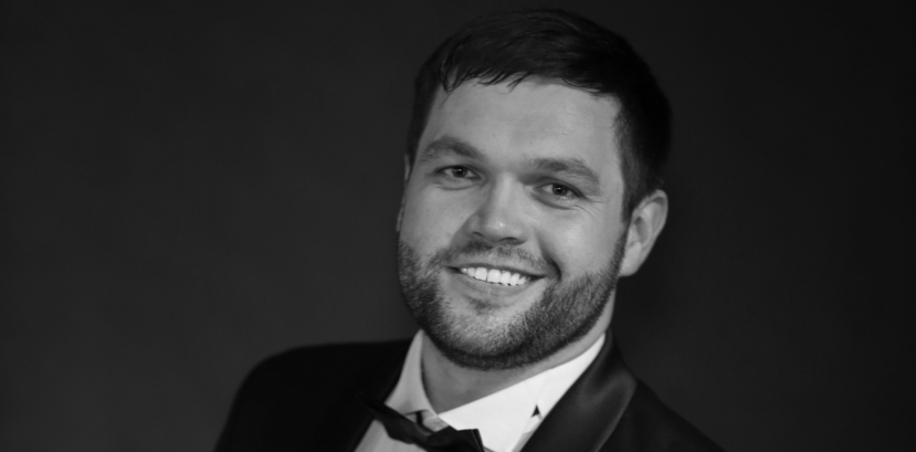 młody mężczyzna w garniturze i muszce uśmiecha się patrząc na wprost, jedna ręka zgięta przytrzymuje marynarkę na poziomie brzucha