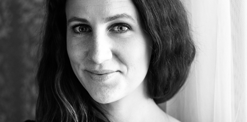 młoda kobieta w długich włosach zebranych swobodnie i położonych na jednym ramieniu patrzy na wprost, lekko się uśmiecha