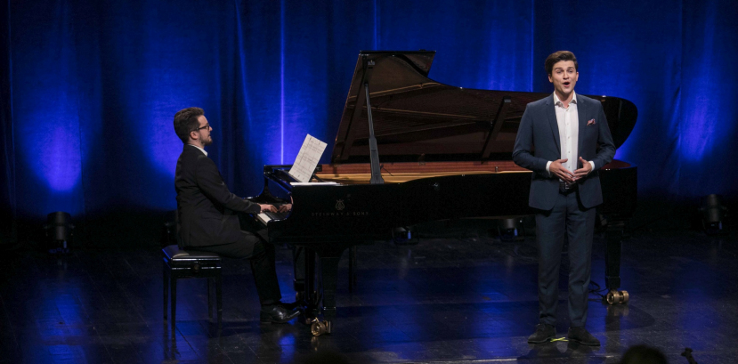 młody mężczyzna stoi na scenie, lekko za nim znajduje się fortepian, na którym gra drugi mężczyzna