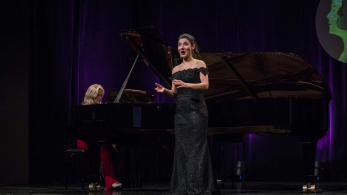 Kobieta w eleganckiej czarnej sukni stoi na scenie. Za ną widać drugą kobietę grającą na fortepianie.